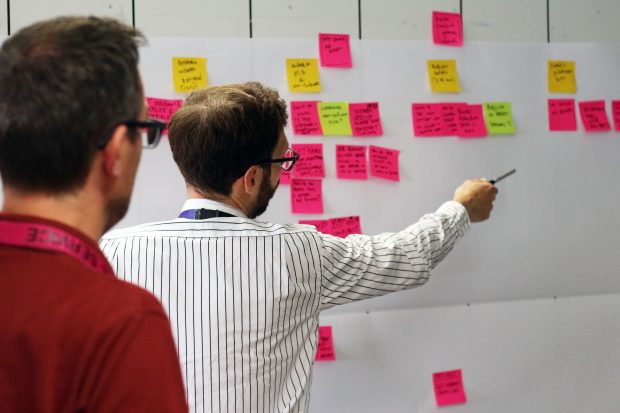 Two people looking at a board with lots of Post It notes attached. One is pointing to one of the notes