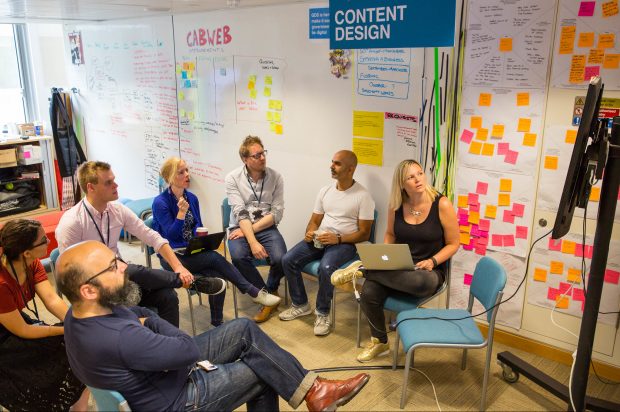 Seven members of a team sat on chairs in front of three large white boards with a lot of post-it notes attached to it. The team are looking at a television screen - one has a laptop on their lap and is presenting to the rest of the team.