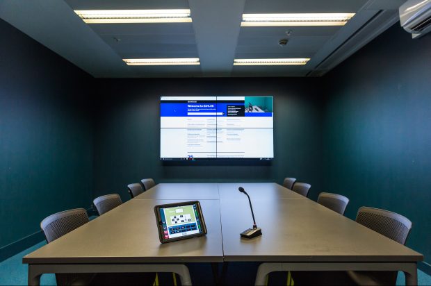 One of the user research labs at GDS. The picture shows a large table in the centre of the room, with a tablet and a microphone on it. At the back of the room is a large television screen, on which the website GOV.UK is displayed.