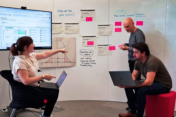 The author and two colleagues at work. They are in front of a wall on which there are bright sticky notes, printed pieces of paper and a large television screen.