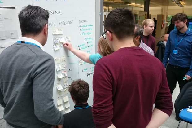 A large team huddled around a white board. There are lots of sticky notes on the board, and a large number of annotations made in black and bright green ink.